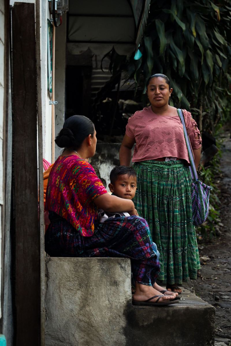 Una madre con traje típico guatemalteco conversa con otra mujer mientras su hijo las escucha